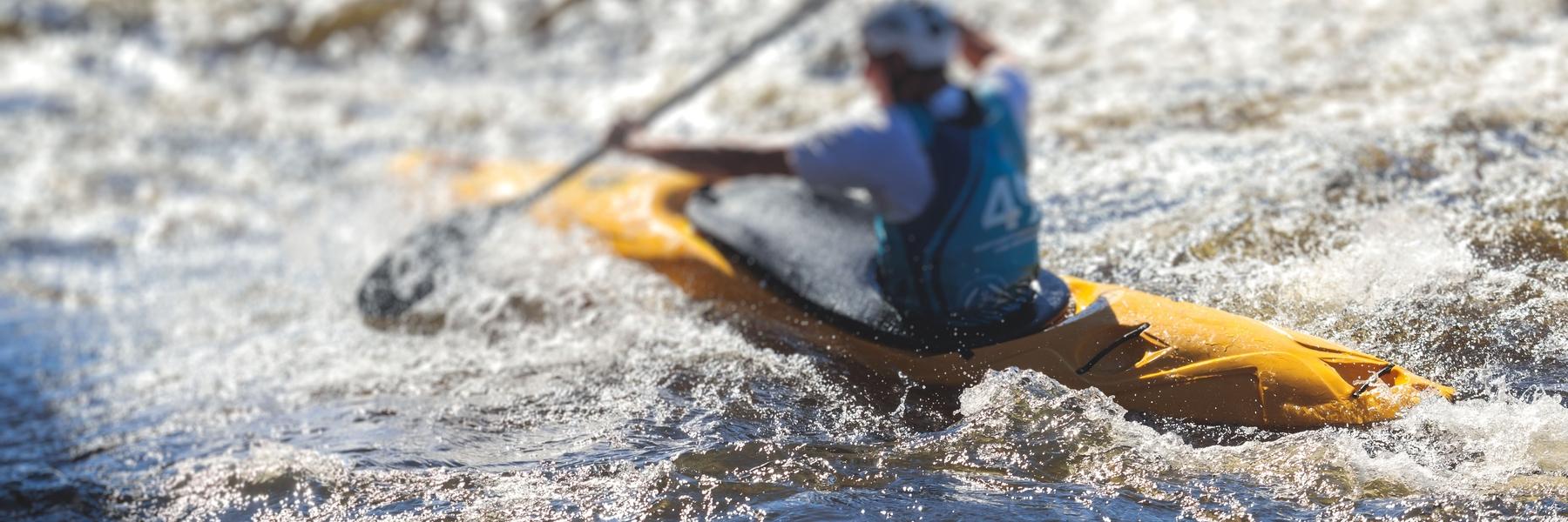Paddler in einem gelben Kajak kämpft sich durch turbulent gewelltes Wasser.