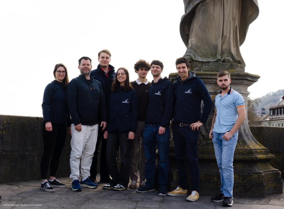 Gruppenfoto von acht Personen vor einer Statue, alle tragen verschiedene Oberbekleidung und stehen auf einer steinernen Fläche.